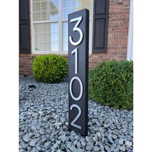 Könnte beinhalten: Schwarzes Hausnummernschild mit silbernen Zahlen 3102. Das Schild steht in einem Bett aus kleinen grauen Steinen.