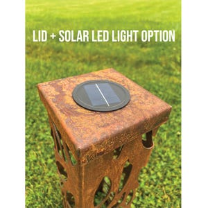 K&ouml;nnte beinhalten: Eine verwitterte, quadratische Metall-Au&szlig;enlaterne mit Solar-LED-Licht oben. Die Laterne hat ausgeschnittene Designs an den Seiten. Der Text "LID + SOLAR LED LIGHT OPTION" ist &uuml;ber der Laterne zu sehen. Der Hintergrund ist gr&uuml;nes Gras.