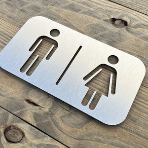 Puede incluir: Un letrero rectangular de baño de metal cepillado con figuras recortadas de hombre y mujer. Las figuras están separadas por una línea vertical. El letrero está montado sobre una superficie de madera.