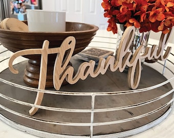 May include: A wooden "Thankful" sign in a cursive font, layered for a 3D effect, sits on a decorative tray. The sign is light brown, and the tray has a distressed white metal rim. A wooden bowl and flowers are in the background.