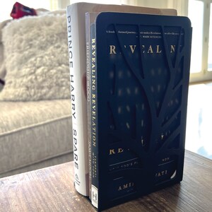 May include: A black bookend with a tree design holds two books. The book on the left is titled "Prince Harry Spare" and the book on the right is titled "Revealing Revelation".