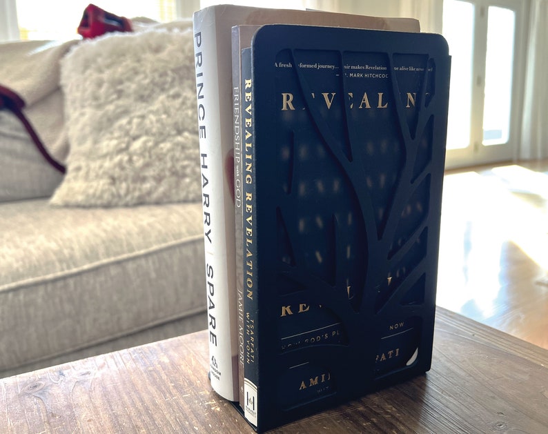 May include: A black bookend with a tree design holds two books. The book on the left is titled "Prince Harry Spare" and the book on the right is titled "Revealing Revelation".