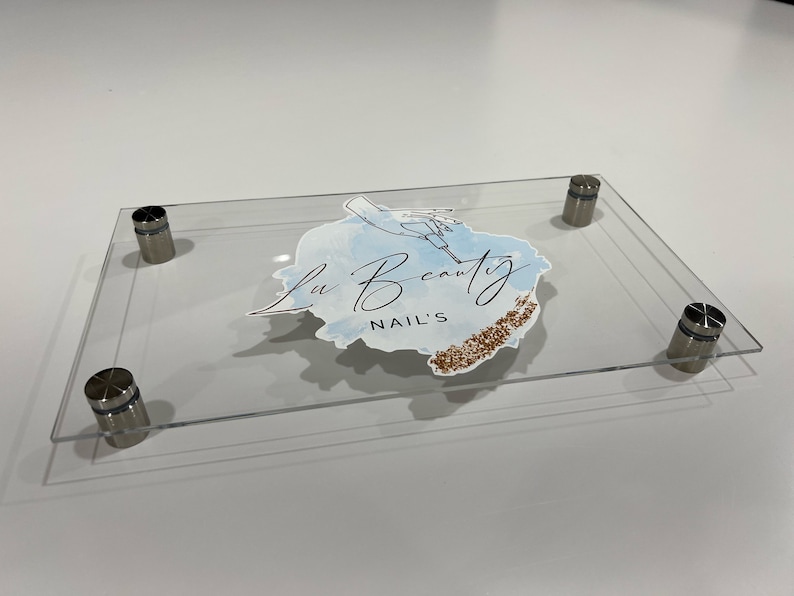 Pu&ograve; includere: Insegna in acrilico trasparente con la scritta "Lu Beauty NAIL'S" in corsivo, con un design ad acquerello e dettagli glitterati dorati. L'insegna &egrave; tenuta in posizione da quattro distanziatori argentati.