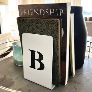 Op de afbeelding: Een witte metalen boekenlegger met de letter "B" uitgesneden in zwart. De boekenlegger houdt een stapel boeken vast, waaronder een met de titel "Friendship".