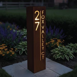 May include: A rectangular, illuminated address sign with the number "27" and the text "FOX VALLEY CT" in a warm, glowing font. The sign is made of a dark, weathered metal and is set in a garden with various plants and flowers.