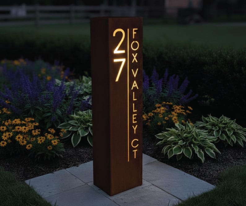 May include: A rectangular, brown metal address sign with the number "27" illuminated in a warm, golden light. The text "FOX VALLEY CT" is vertically displayed. The sign is set in a garden with flowers and greenery.
