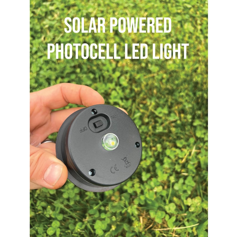 K&ouml;nnte beinhalten: Ein schwarzes Solar-LED-Fotozellenlicht mit dem Text "SOLAR POWERED PHOTOCELL LED LIGHT". Das Licht hat einen Ein-/Ausschalter und eine kleine, helle LED. Der Hintergrund ist gr&uuml;nes Gras.