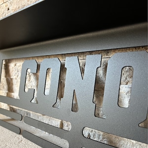 Puede incluir: Un expositor de medallas de metal negro montado en la pared con la palabra "CONO" recortada en el centro. El expositor tiene un estante en la parte superior y una serie de barras horizontales para colgar medallas.