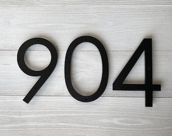 Puede incluir: Números de casa 904 de metal negro. Los números están fijados a una superficie de madera blanca.