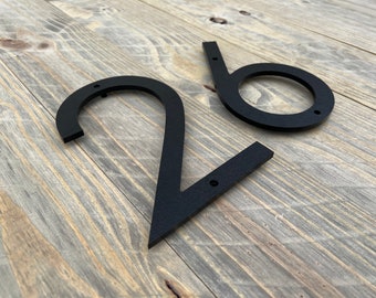 May include: Black metal house numbers, 2 and 6, with a modern, minimalist design. The numbers are mounted on a wooden surface.