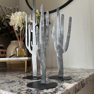 以下が含まれることがあります： 銀色の仕上げの金属製のサボテンの彫刻が3つあります。彫刻は、斑点のある模様の白いカウンタートップの上に置かれています。