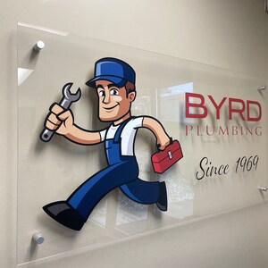 Pu&ograve; includere: Insegna in acrilico trasparente con un idraulico dei cartoni animati in tuta blu, con una chiave inglese e una cassetta degli attrezzi. L'insegna recita "BYRD PLUMBING" in testo rosso e nero, con "Since 1969" sotto. L'insegna &egrave; montata su una parete beige.