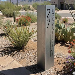 Może przedstawiać: Prostokątny, srebrny metalowy znak adresowy z numerem "27" i czarnym napisem "FOX VALLEY CT". Znak znajduje się na zewnątrz, otoczony pustynnym krajobrazem z kaktusami i kwitnącymi roślinami.