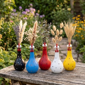 Peut inclure: Cinq vases en céramique en forme de poules, chacun d'une couleur distincte : noir, bleu, rouge, blanc et jaune. Chaque vase possède une crête rouge, des yeux blancs et un bec, et est rempli de diverses fleurs séchées, herbes et épis de blé.