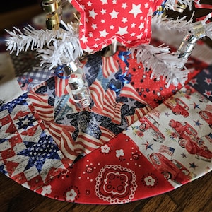 May include: A red, white, and blue patchwork fabric tree skirt with a red star-shaped ornament. The skirt is decorated with stars, stripes, and red trucks.