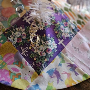 May include: A patchwork fabric Easter tree skirt with a purple floral design, a white feather trim, and a silver ribbon. The skirt features a variety of Easter-themed patterns, including bunnies, eggs, and crosses.