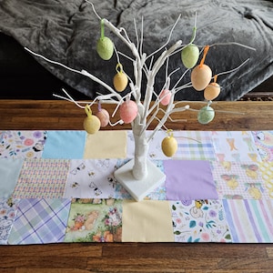 May include: A white twig tree with pastel colored Easter eggs hanging from the branches. The tree is on a patchwork table runner with a variety of floral and geometric patterns in shades of yellow, blue, and purple.