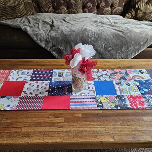 Könnte beinhalten: Ein roter, weißer und blauer Patchwork-Tischläufer mit verschiedenen Comicfiguren und Mustern. Eine Glasvase mit roten, weißen und rosa Blumen steht auf dem Tischläufer.