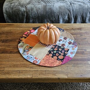 Op de afbeelding: Een ronde patchwork placemat met herfstthema stoffen met pompoen- en bloemmotieven. Een decoratieve, oranje pompoen ligt op de placemat op een houten tafel. De tafel heeft een donkere houten afwerking.