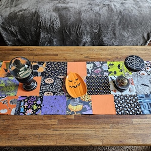 May include: A Halloween-themed table runner with a patchwork design featuring pumpkins, bats, and other spooky motifs. Decorative items include a crystal ball, a jack-o'-lantern shaped dish, and a coaster with a spiderweb design.