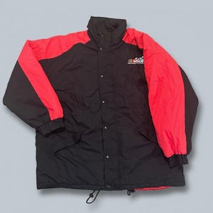 Peut inclure: Une veste noire et rouge à blocs de couleurs avec un col montant, des manches longues à poignets élastiques et une fermeture à boutons-pression. La poitrine arbore un logo NASCAR blanc avec un drapeau à damier. L'ourlet possède un cordon de serrage.