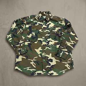 May include: A long-sleeve button-down shirt featuring a camouflage pattern in shades of olive green, dark brown, black, and tan. The shirt includes a standard collar, two chest pockets with buttoned flaps, and buttoned cuffs on the sleeves.