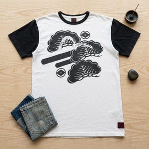 Pode incluir: Uma t-shirt de manga curta com painel frontal branco, mangas pretas e gola redonda preta. A frente apresenta uma impressão gráfica preta de três ramos de pinheiro estilizados e dois pequenos emblemas florais circulares. Uma pequena etiqueta vermelha encontra-se na bainha.