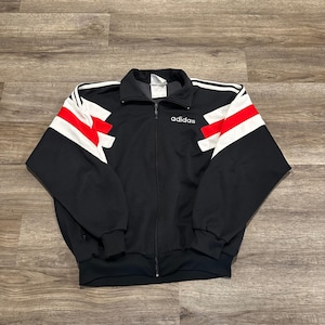 May include: Black Adidas track jacket with white and red detailing on the sleeves. The jacket features a full-length zip and the Adidas logo in white on the chest. The jacket is made of a smooth, athletic material.