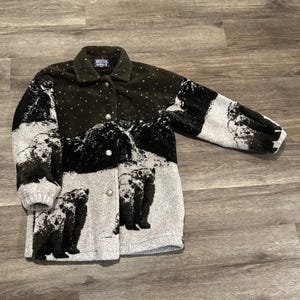 May include: A button-down fleece jacket with a mountain and bear design. The jacket features a black and white landscape with bears, and the text "ARCTIC CIRCLE" on the collar. The jacket has a dark green background with white dots.