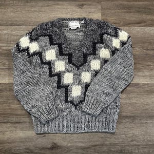 Könnte beinhalten: Ein grauer, weißer und schwarzer V-Ausschnitt-Pullover mit geometrischem Muster. Der Pullover hat ein strukturiertes Strickdesign mit einem sich wiederholenden Muster aus weißen Quadraten und schwarzen Linien. Die Ärmel und der Körper sind eine Mischung aus grauem und weißem Garn.