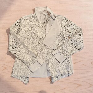 May include: A beige lace blouse with long sleeves and a bow tie collar. The blouse features a delicate floral lace overlay, with a solid beige fabric underneath. The cuffs are also solid beige. The blouse is laid flat on a light pink surface.