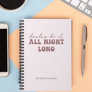 May include: A white spiral bound notebook with the text "doulas do it ALL NIGHT LONG" in pink lettering. The notebook is on a light orange surface with a silver pen and a white keyboard. The text "My Doula Log Book" is at the bottom of the notebook.