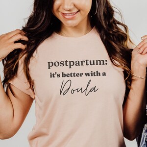 May include: A woman wearing a heather peach t-shirt with the text "postpartum: it's better with a Doula" printed on it. She is holding a denim jacket in her right hand.