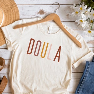 May include: A beige t-shirt with the word "DOULA" printed in brown, orange, pink, and red letters. The shirt is hanging on a wooden hanger and is displayed on a white wooden background.