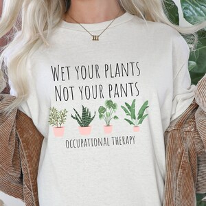 May include: A white long-sleeved t-shirt with the text "Wet your plants not your pants" and a row of potted plants below. The text "Occupational Therapy" is printed below the plants.