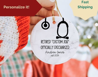 May include: White marble Christmas bauble with a black stethoscope design and the text "Retired "Custom Job" Officially Discharged" with a name and year.
