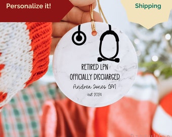 May include: White marble Christmas bauble with a black stethoscope and the text "Retired LPN Officially Discharged Andrea Jones LPN est 2024".