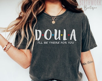 May include: A dark gray t-shirt with the text "DOULA I'LL BE THERE FOR YOU" in white with colorful dots. The shirt is worn by a person with long blonde hair and blue jeans.