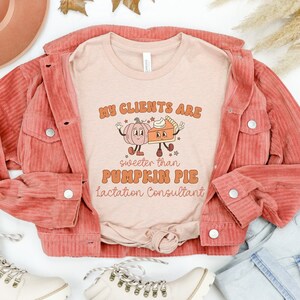 May include: A pink t-shirt with the text "My clients are sweeter than pumpkin pie Lactation Consultant" and a cartoon pumpkin and pie slice. The shirt is laid out on a white surface with a denim jacket, jeans, and boots.