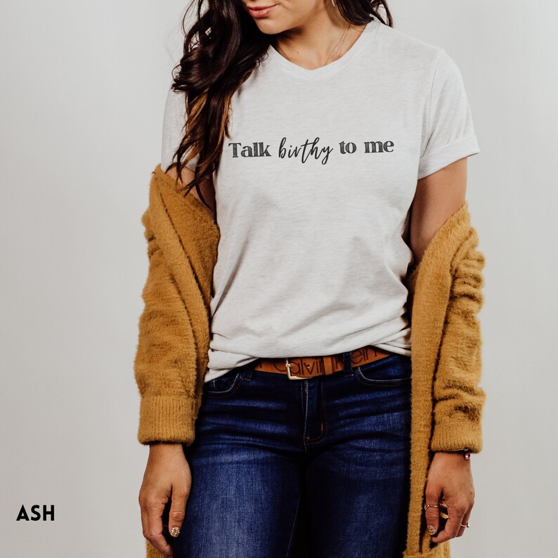 Puede incluir: Una camiseta blanca con el texto "Talk birthy to me" impreso en la parte delantera. La camiseta la lleva puesta una persona con el pelo largo y casta&ntilde;o y un c&aacute;rdigan marr&oacute;n.