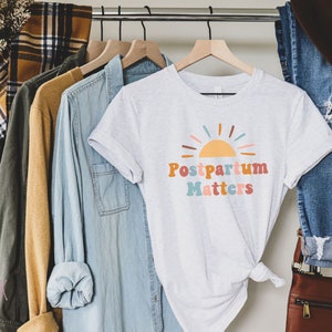 May include: A white t-shirt with a colorful graphic of a sun and the text "Postpartum Matters".