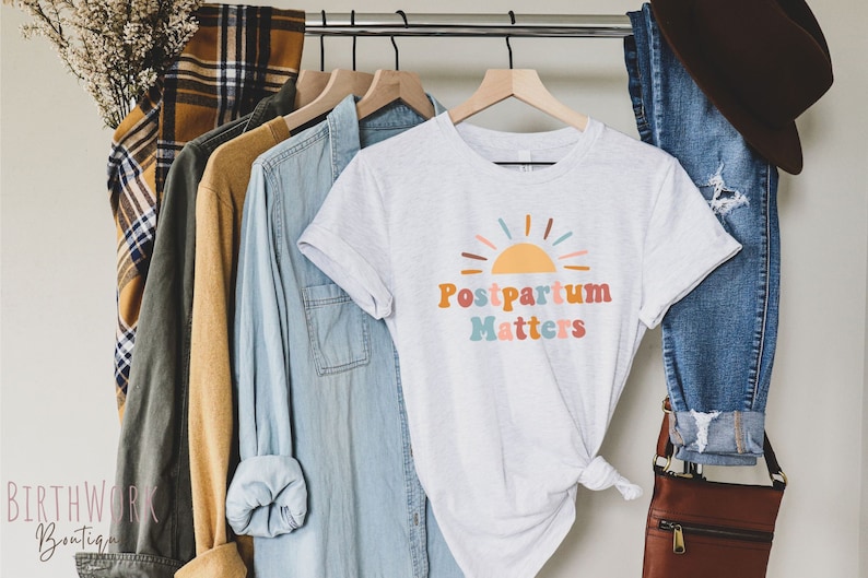 May include: A white t-shirt with a colorful graphic of a sun and the text "Postpartum Matters".