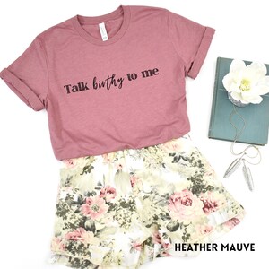 Puede incluir: Una camiseta de color malva con el texto "Talk birthy to me" impreso en negro. La camiseta se muestra sobre un fondo blanco con unos pantalones cortos florales y un libro.