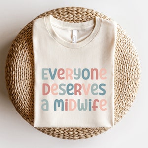 May include: A beige t-shirt with the text "Everyone deserves a midwife" in colorful letters. The letters are in shades of pink, blue, and green.