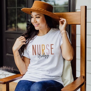 May include: A white t-shirt with the text "NURSE" in brown, yellow, and pink lettering. The text "Urology" is written in cursive below the word "NURSE".