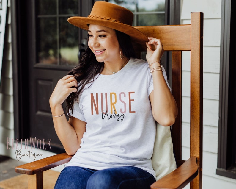 May include: A white t-shirt with the text "NURSE" in brown, yellow, and pink lettering. The text "Urology" is written in cursive below the word "NURSE".