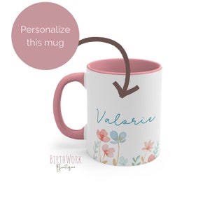 May include: A pink and white ceramic mug with a pink handle. The mug has a floral design with blue and pink flowers and green leaves. The name "Valorie" is written in blue on the mug.