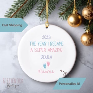 May include: White ceramic ornament with the text "2023 The Year I Became A Super Amazing Doula" and a pink and blue footprint graphic. The text "Naomi" is written below the footprint graphic. The ornament has a gold string for hanging.