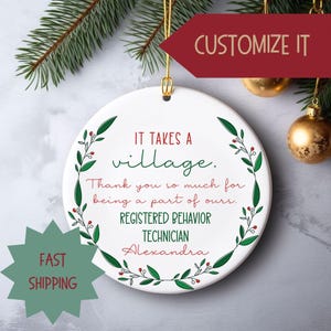 May include: White round ornament with a green wreath border and red berries. The ornament says "It takes a village. Thank you so much for being a part of ours. Registered Behavior Technician Alexandra".