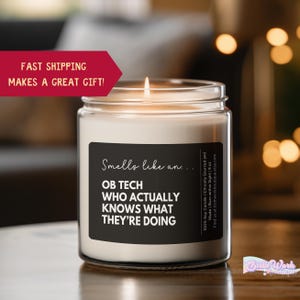 May include: A white candle with a black label that reads "Smells like an... OB TECH WHO ACTUALLY KNOWS WHAT THEY'RE DOING". The candle is lit and has a warm glow.